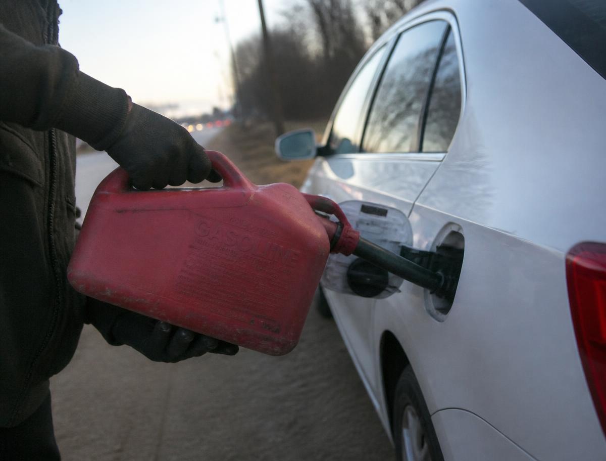 Gloved hands pouring fuel from a red gas can into a car's filler neck on a roadside shoulder